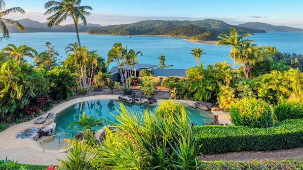 A pool surrounded by tropical vegetation and a view of a large body of water with mountains in the background.