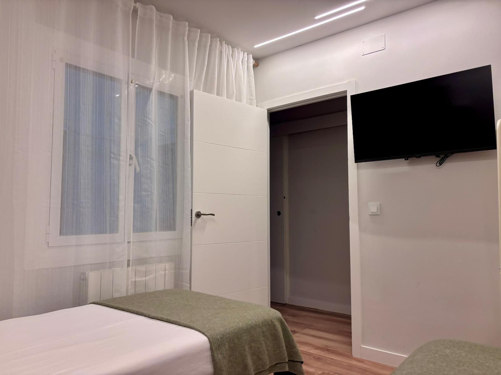 A bedroom with a bed, a green blanket, a white curtain, a television mounted on the wall, and a wooden floor.