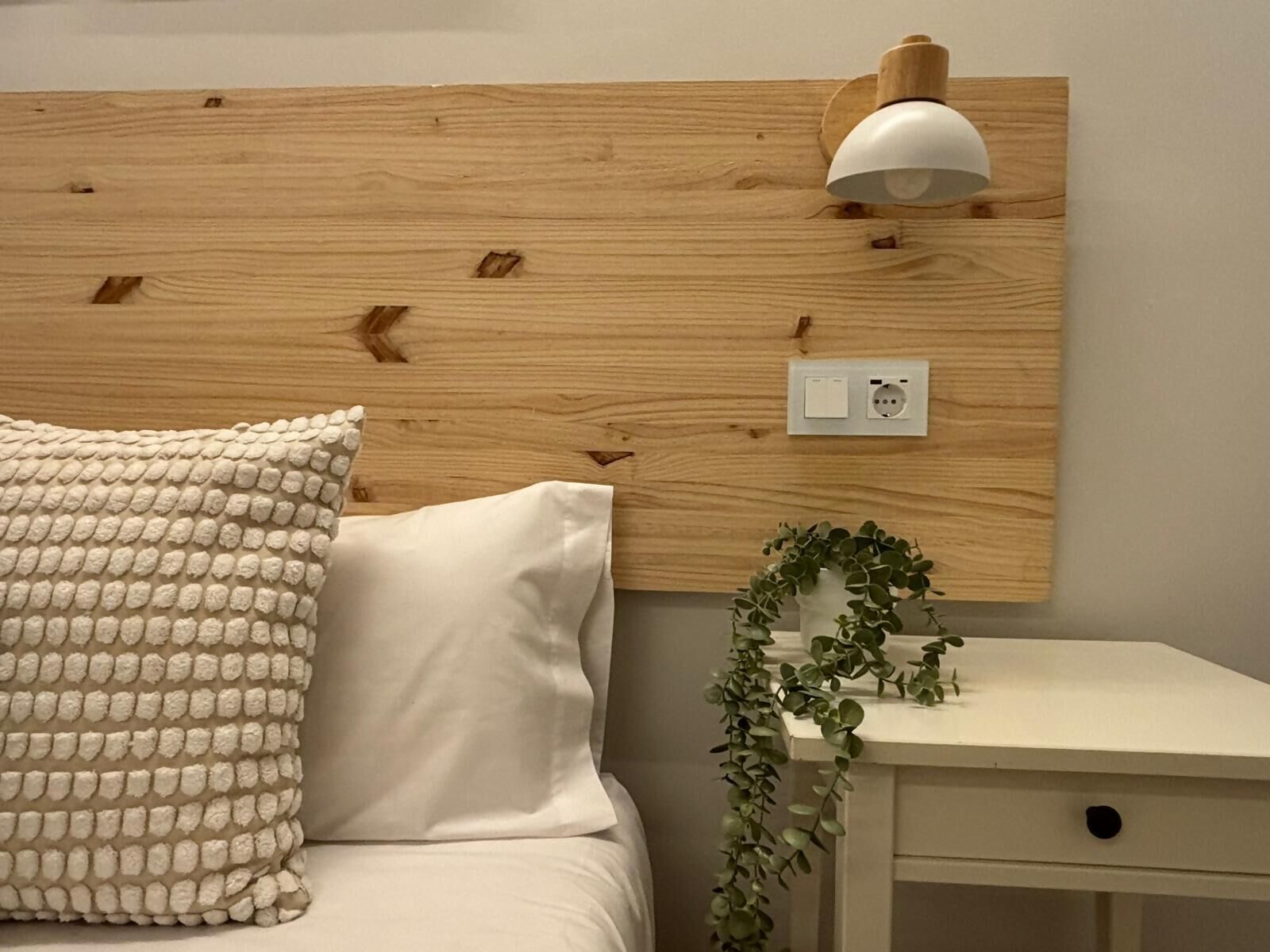 A bedroom with a wooden headboard, a bedside lamp, a white bedside table with a plant, and a textured pillow.