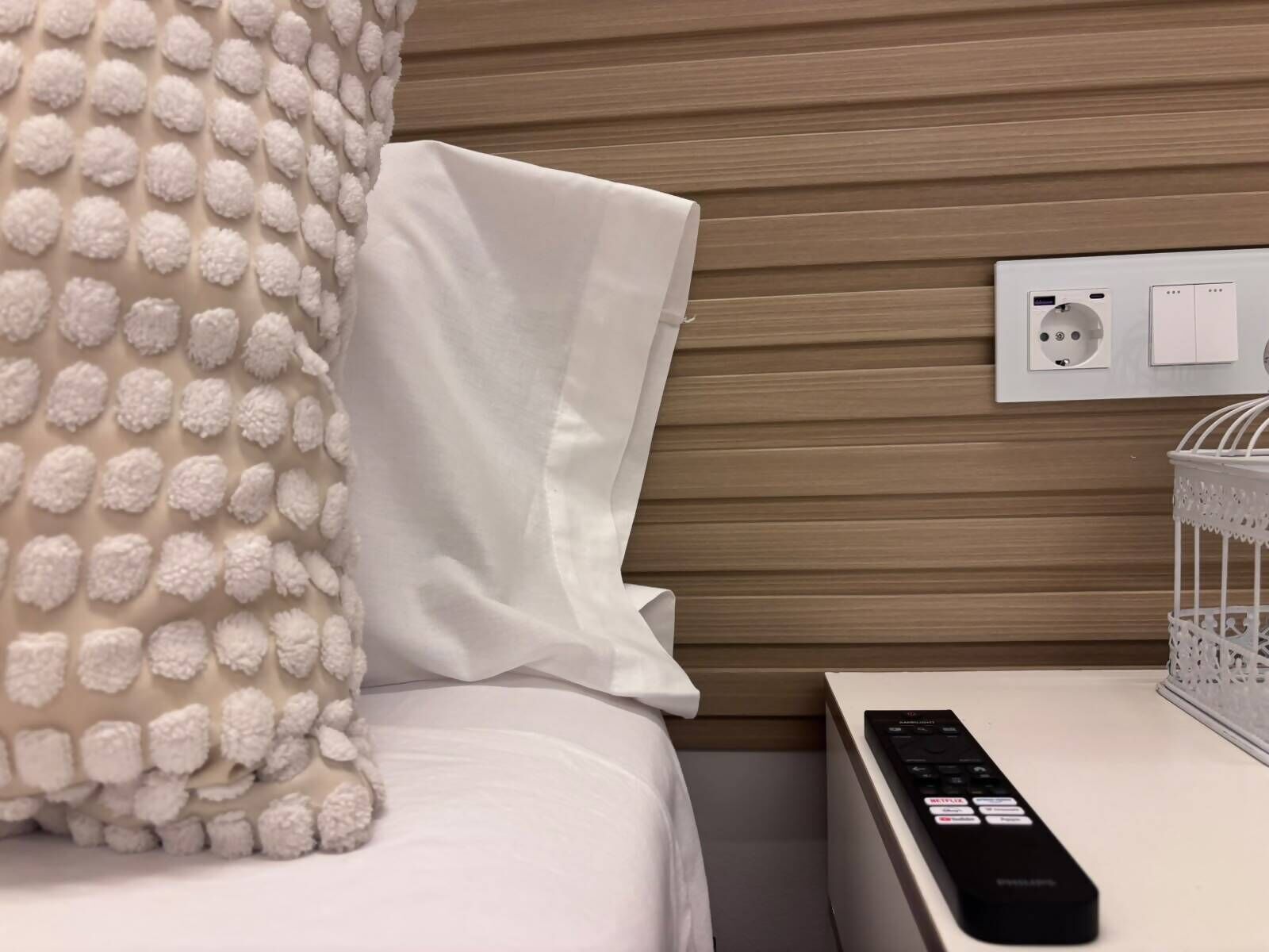 Una cama bien hecha con un almohadón blanco y una colcha texturizada, un control remoto sobre una mesita de noche y un enchufe con toma de corriente.