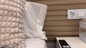 Una cama bien hecha con un almohadón blanco y una colcha texturizada, un control remoto sobre una mesita de noche y un enchufe con toma de corriente.