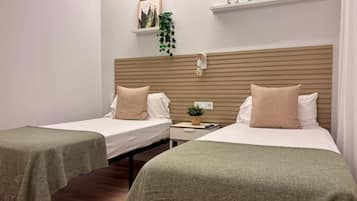 A hotel room with two single beds, a bedside table with a lamp, a wall-mounted shelf with decorative items, and a potted plant.