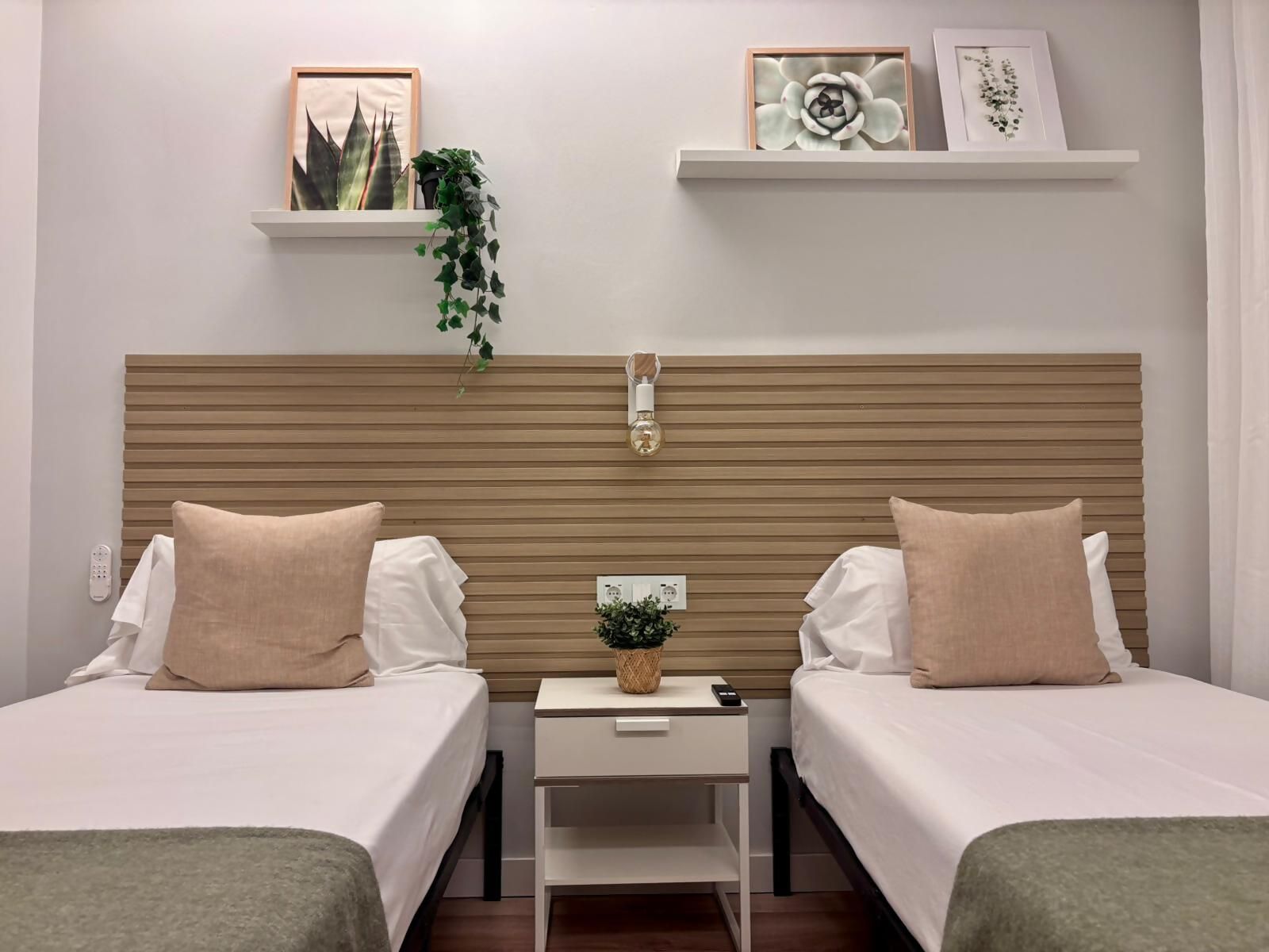 Habitación de hotel con dos camas, una mesita de noche con un control remoto, una planta pequeña y estanterías con marcos.