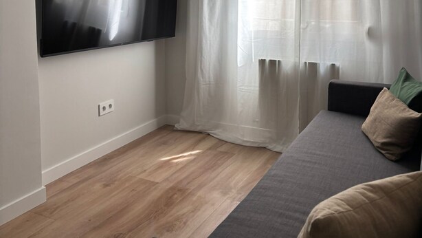 En moderne stue med et fladskærms-tv på væggen, en sofa med puder og lette gardiner ved vinduet.
