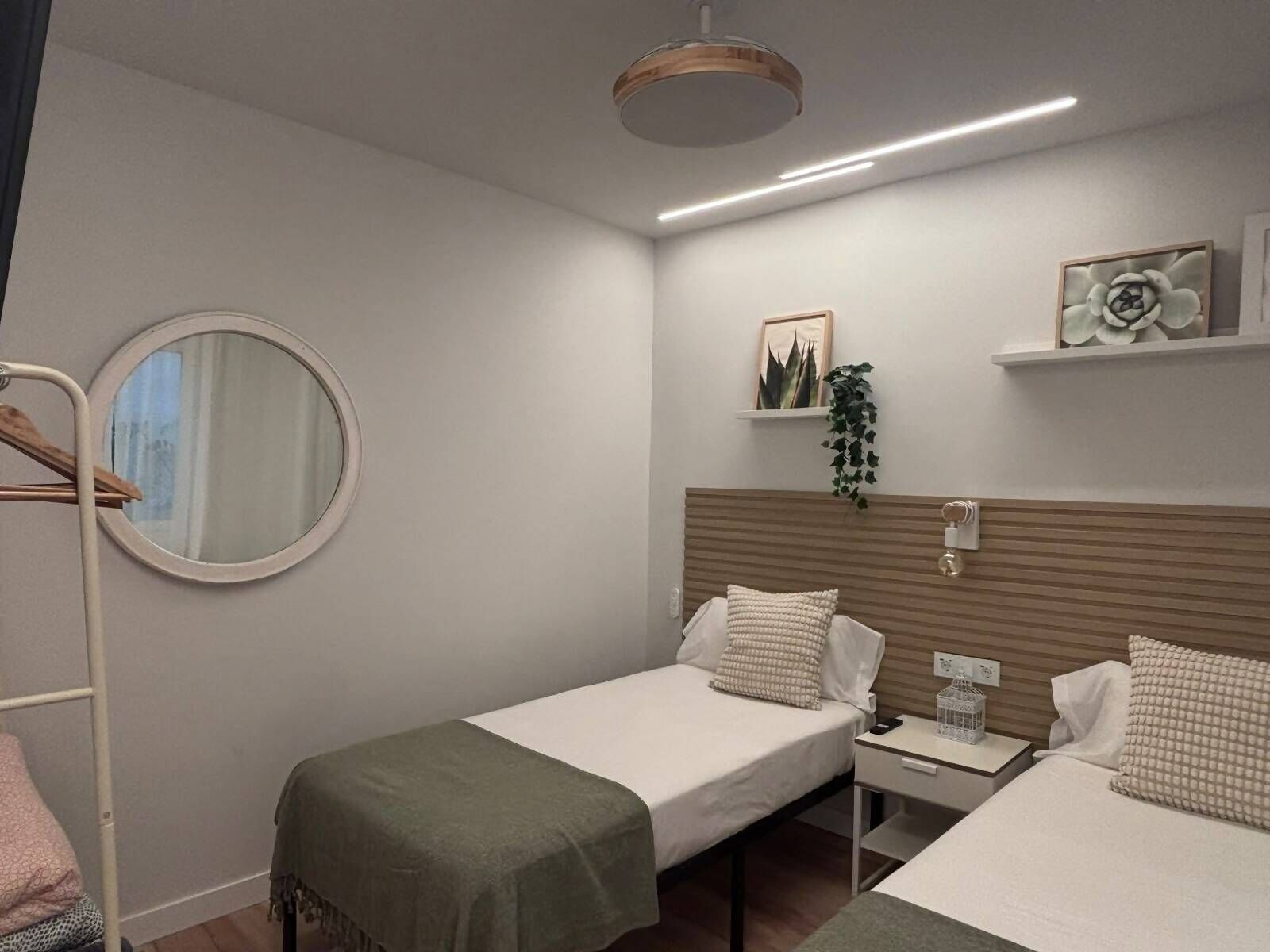 A hotel room with two single beds, a round mirror, a bedside table with a lamp, a shelf with decorative items, and a hanging plant.