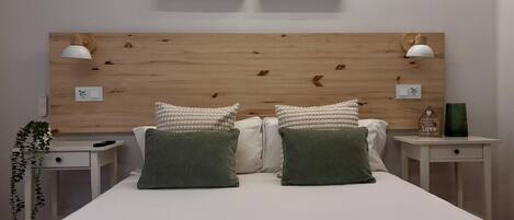 A bedroom with a wooden headboard, two bedside tables, a bed with pillows, a wall-mounted light fixture, and a small plant.