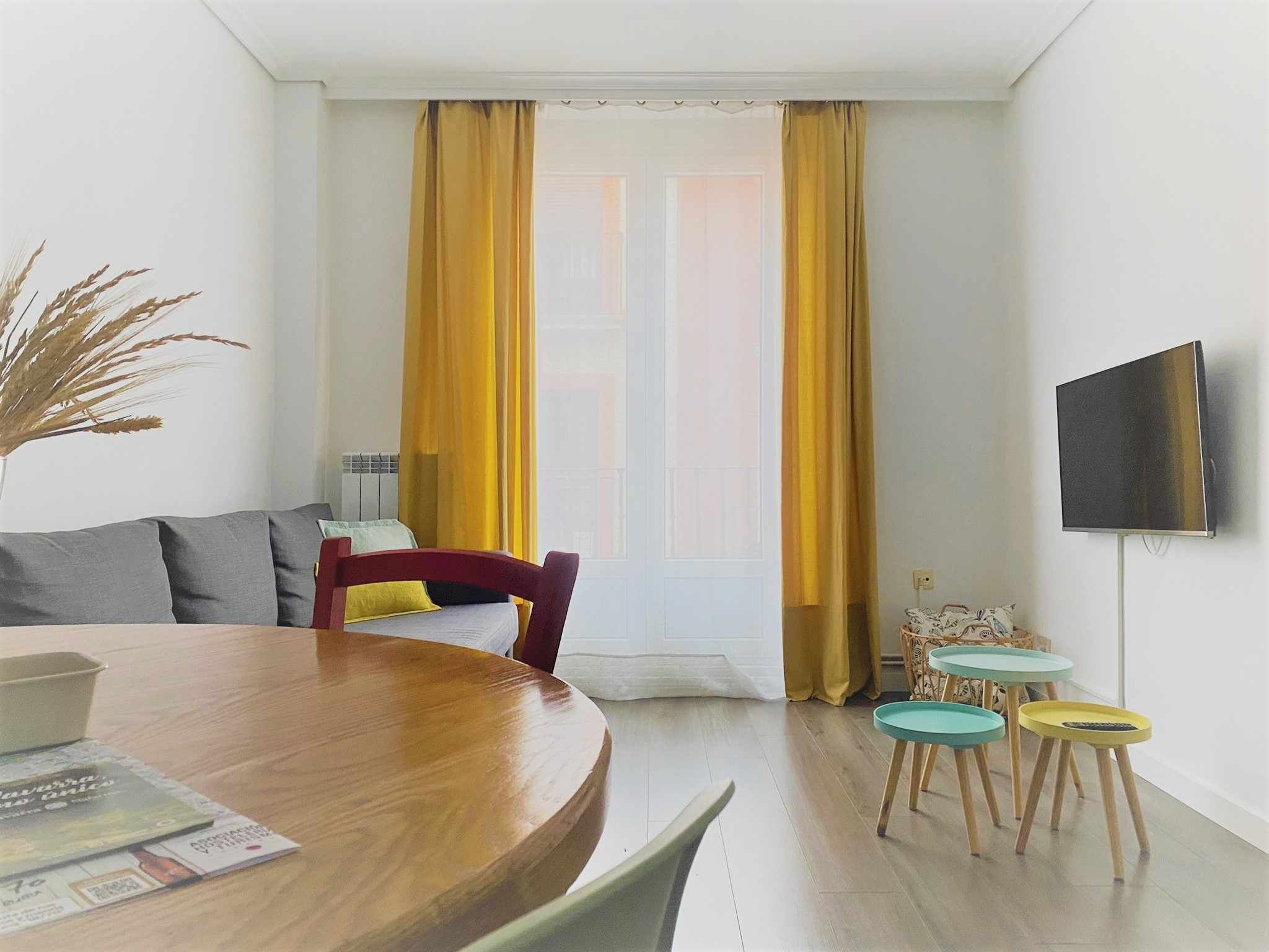 A modern living room with a wooden dining table, a sofa, and a TV mounted on the wall. - Apartamento en el Casco Antiguo de Pamplona - Ayla Camino de Santiago (Pamplona/Iruña)