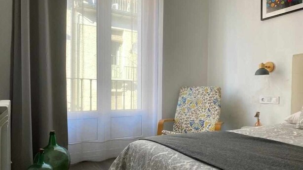 A bedroom with a bed, a chair, a lamp, and a window with a view of buildings. - Apartamento en el Casco Antiguo de Pamplona - Ayla Camino de Santiago (Pamplona/Iruña)