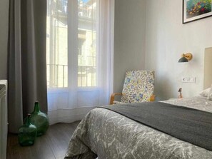 A bedroom with a bed, a chair, a lamp, and a window with a view of buildings. - Apartamento en el Casco Antiguo de Pamplona - Ayla Camino de Santiago (Pamplona/Iruña)