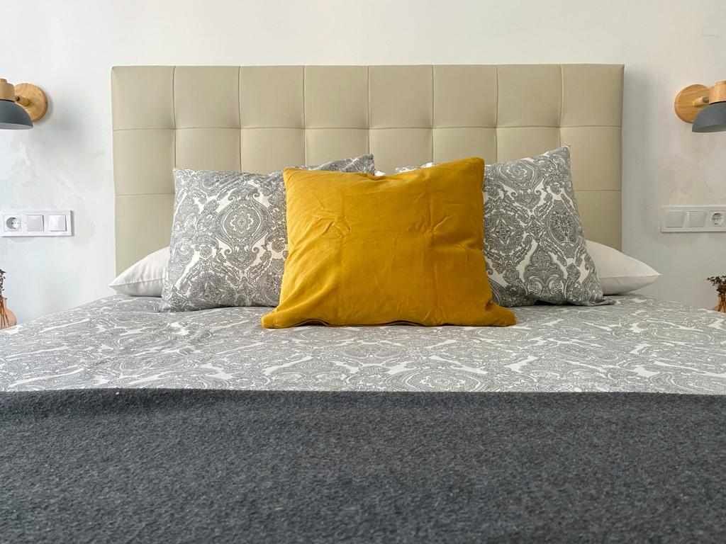 Una cama con cabecera capitonada, dos almohadas estampadas y una manta gris.