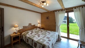 A bedroom with a wooden headboard, a quilted bedspread, bedside lamps, and a view of a green lawn through a glass door. - Spacious 3-Bed Apt for 8, 80m from Cernix Slopes, Pet-Friendly, Terrace, Parking (Crest-Voland)