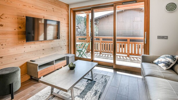 Una sala moderna con una pared de madera, un televisor de pantalla plana, un sofá, una mesa de centro y un ventanal corredizo que da a una terraza exterior.