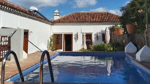 Un área de piscina con solárium de baldosas, un edificio blanco con tejado de tejas rojas y un cielo azul despejado.