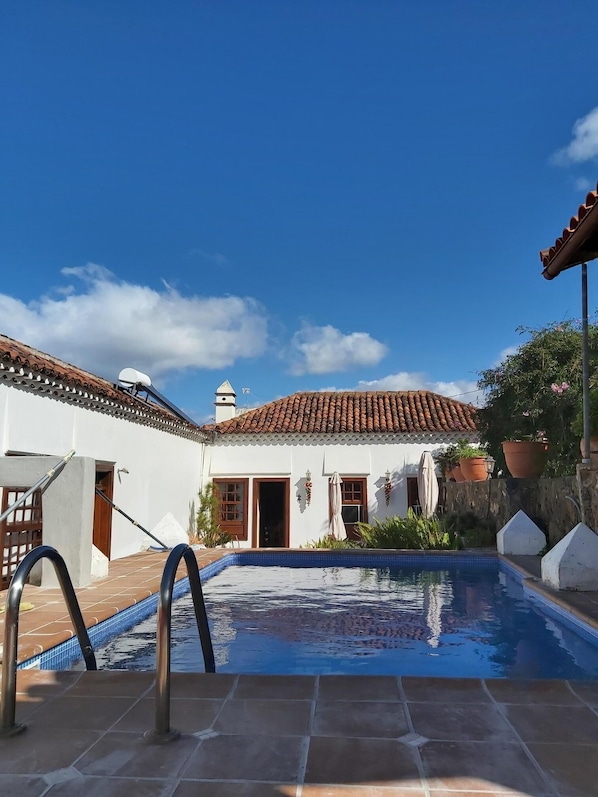 Un área de piscina con solárium de baldosas, un edificio blanco con tejado de tejas rojas y un cielo azul despejado.