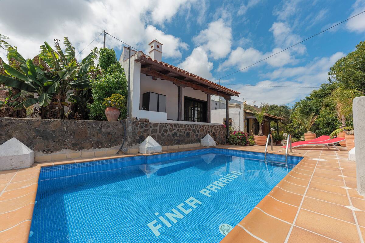 A swimming pool with "FINCA PARIS" written on the tiles, surrounded by a stone wall and a house with a tiled roof.