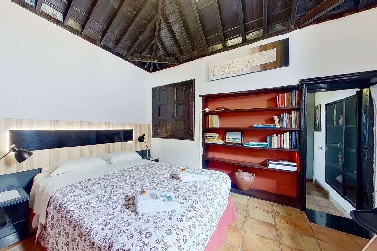 A bedroom with a wooden headboard, a bed with a floral bedspread, a bedside table with a lamp, and a bookshelf filled with books.