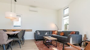 A modern living room with a sofa, coffee table, and chairs. There is a dining area with a table and chairs. The room has large windows and a ceiling fan. - Chalet with wide, deep backyard, sheltered and situated on the southwest<br>Decorated in a contemporary (Burgh-Haamstede)