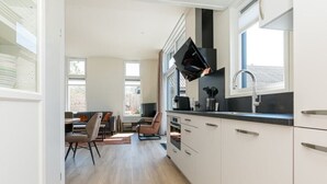 A modern kitchen with white cabinets, a black countertop, and a built-in oven. There is a dining area with a table and chairs, and a window providing natural light. - Chalet with wide, deep backyard, sheltered and situated on the southwest<br>Decorated in a contemporary (Burgh-Haamstede)
