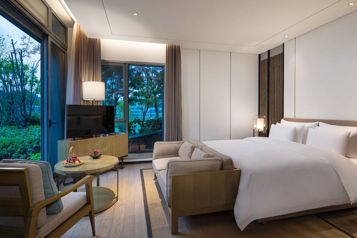 A modern hotel room with a large bed, a TV, a sofa, a dining table, and a view of greenery outside.