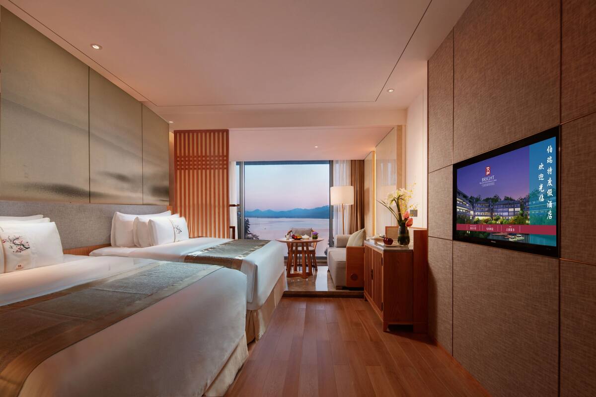 A hotel room with two beds, a TV, a view of the sea, and a wooden cabinet.