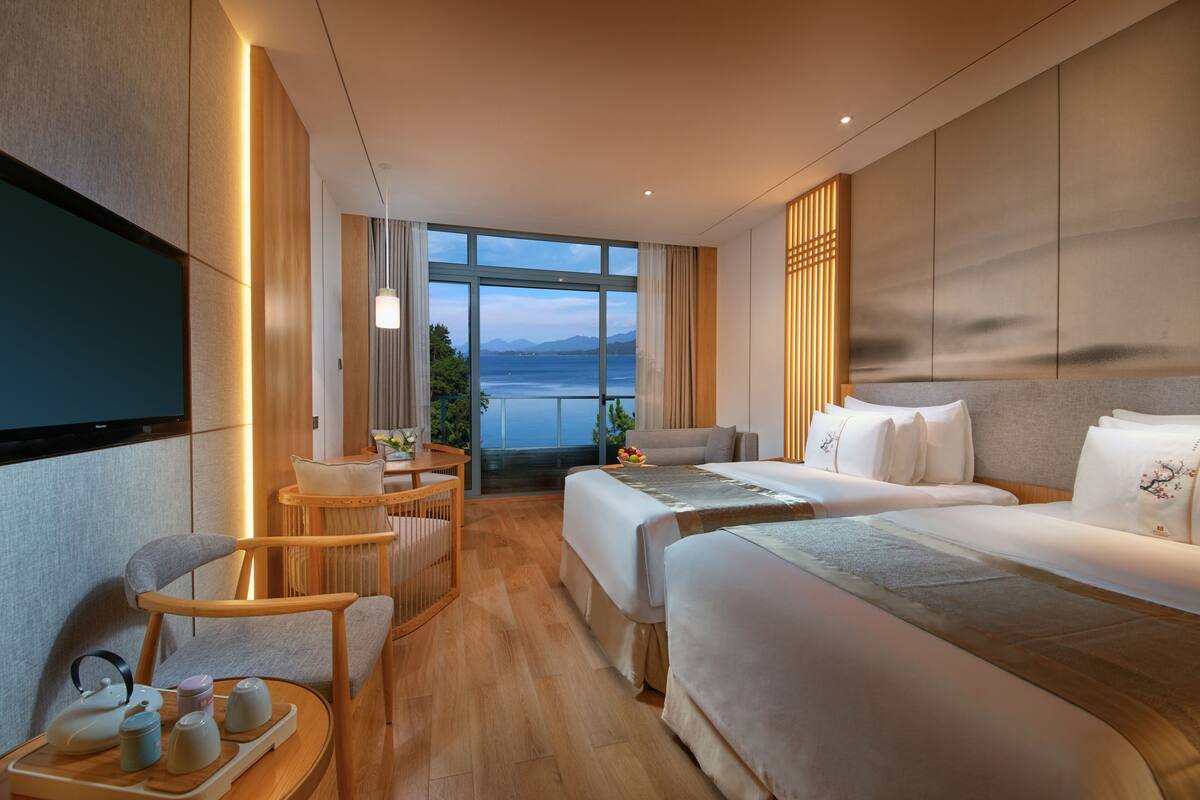 A modern hotel room with a large window offering a view of the sea and mountains. It includes two beds, a sofa, a dining area, and a TV.