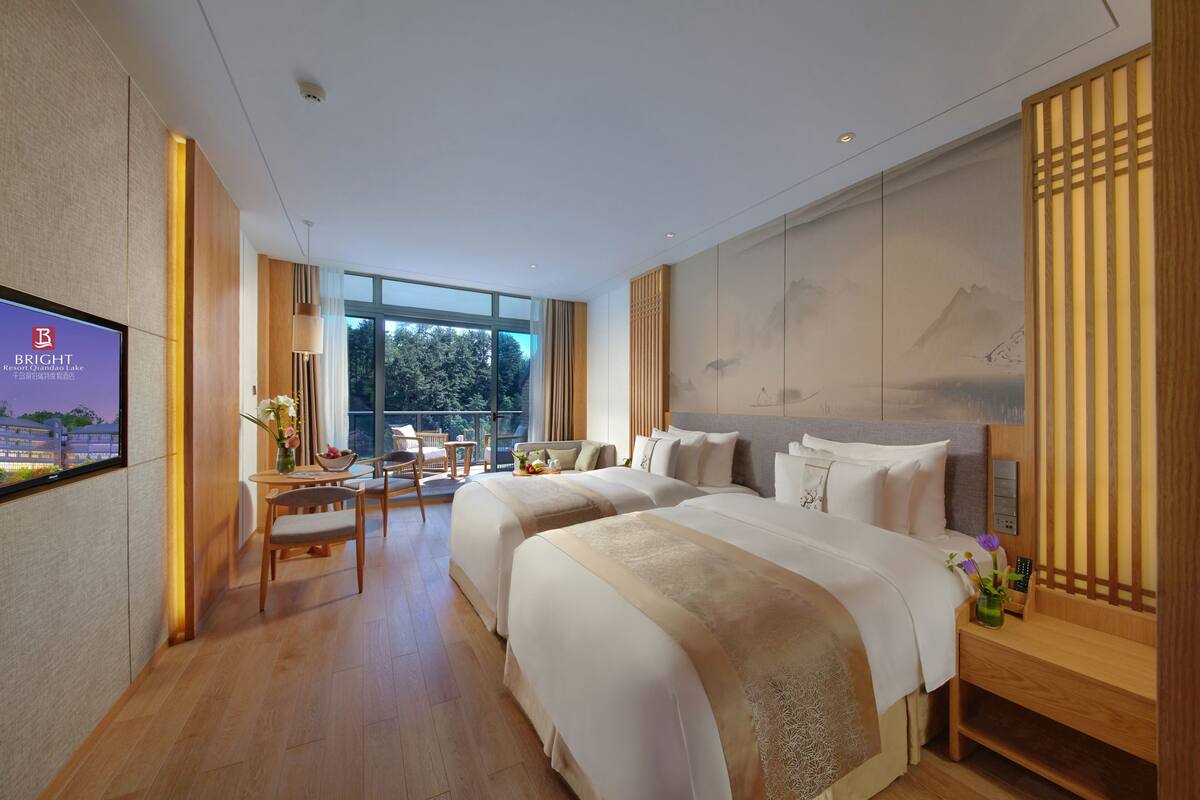 A hotel room with two beds, a TV, a desk with a chair, and a view of the outdoors.