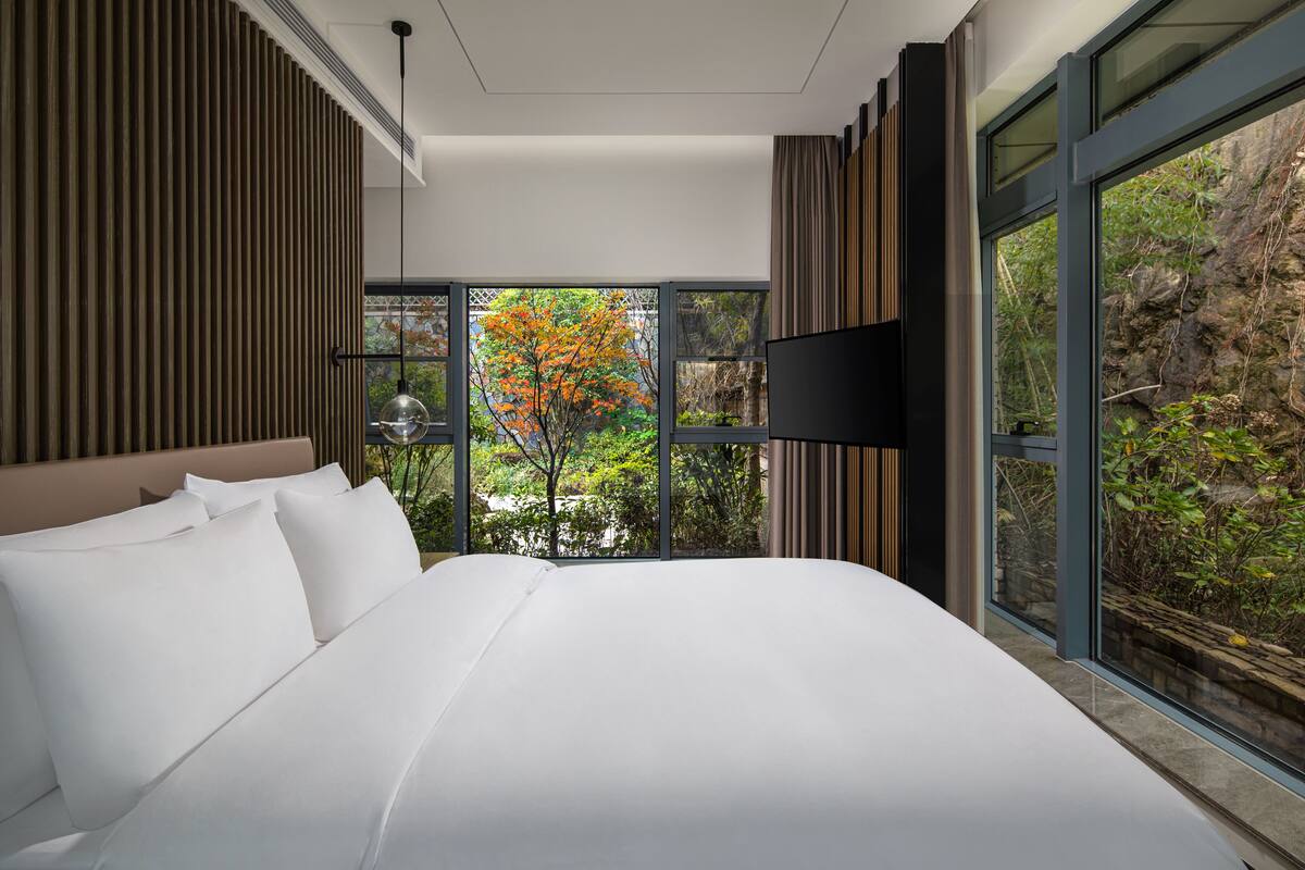 A modern hotel room with a large bed, a flat-screen TV, and a view of a tree through large windows.