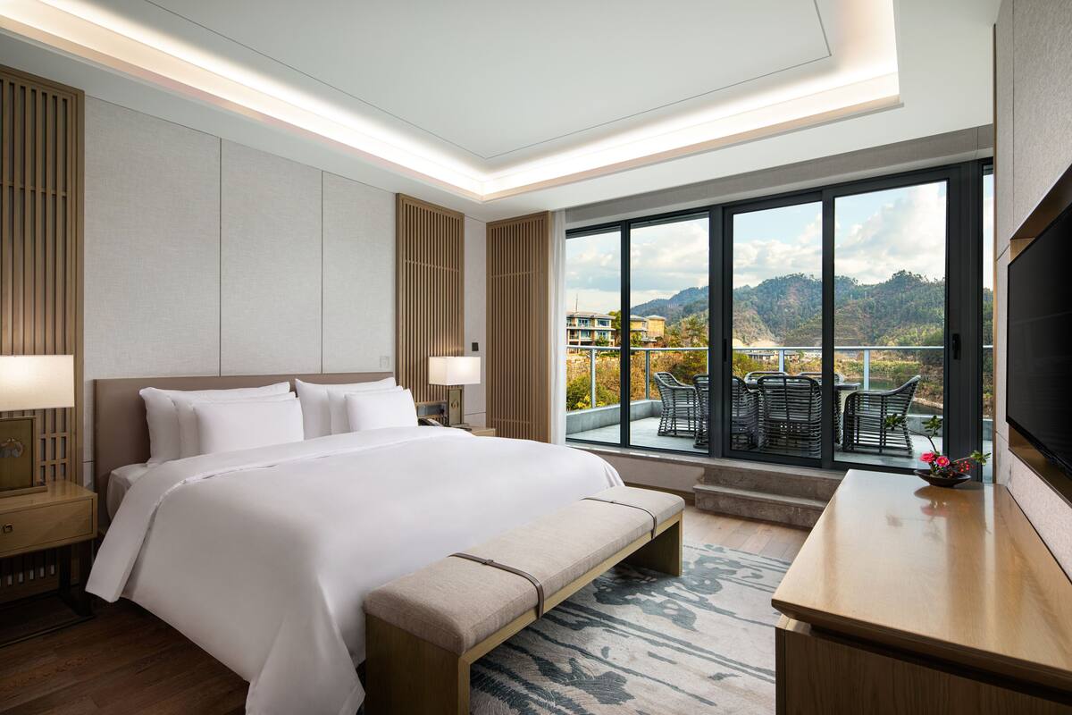 A modern hotel room with a large bed, a bench, a desk, and a view of the outdoors.