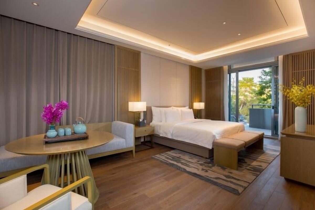 A modern hotel room with a large bed, a seating area with a round table, and a view of the outdoors.