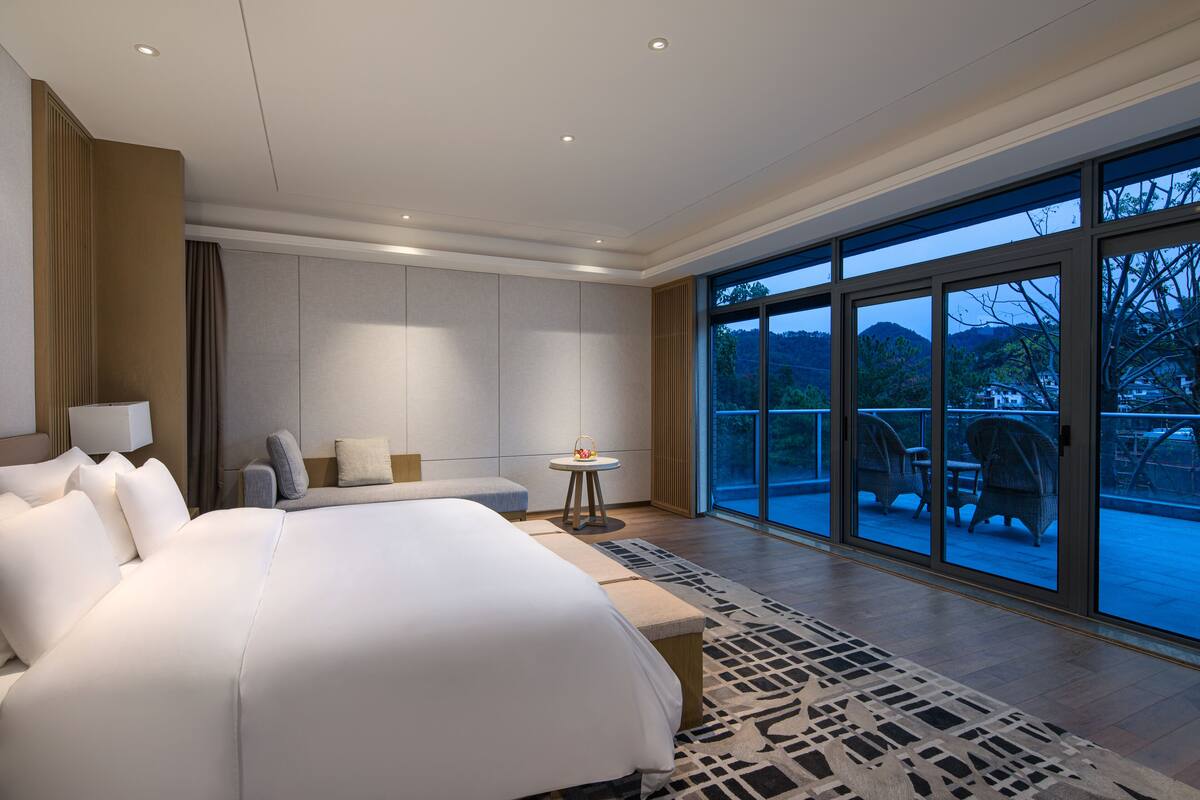 A spacious bedroom with a large bed, a small table, and a chair. There is a balcony with outdoor furniture and a view of the mountains.