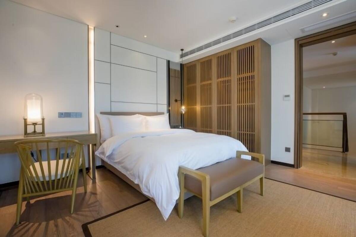 A modern hotel room with a large bed, a desk with a chair, a bedside table, and a sliding wooden door.