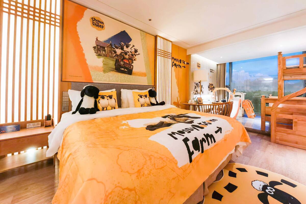 A hotel room with a bed featuring a 'Shaun the Sheep' themed bedspread, a wooden desk, a chair, and a view of the city through large windows.