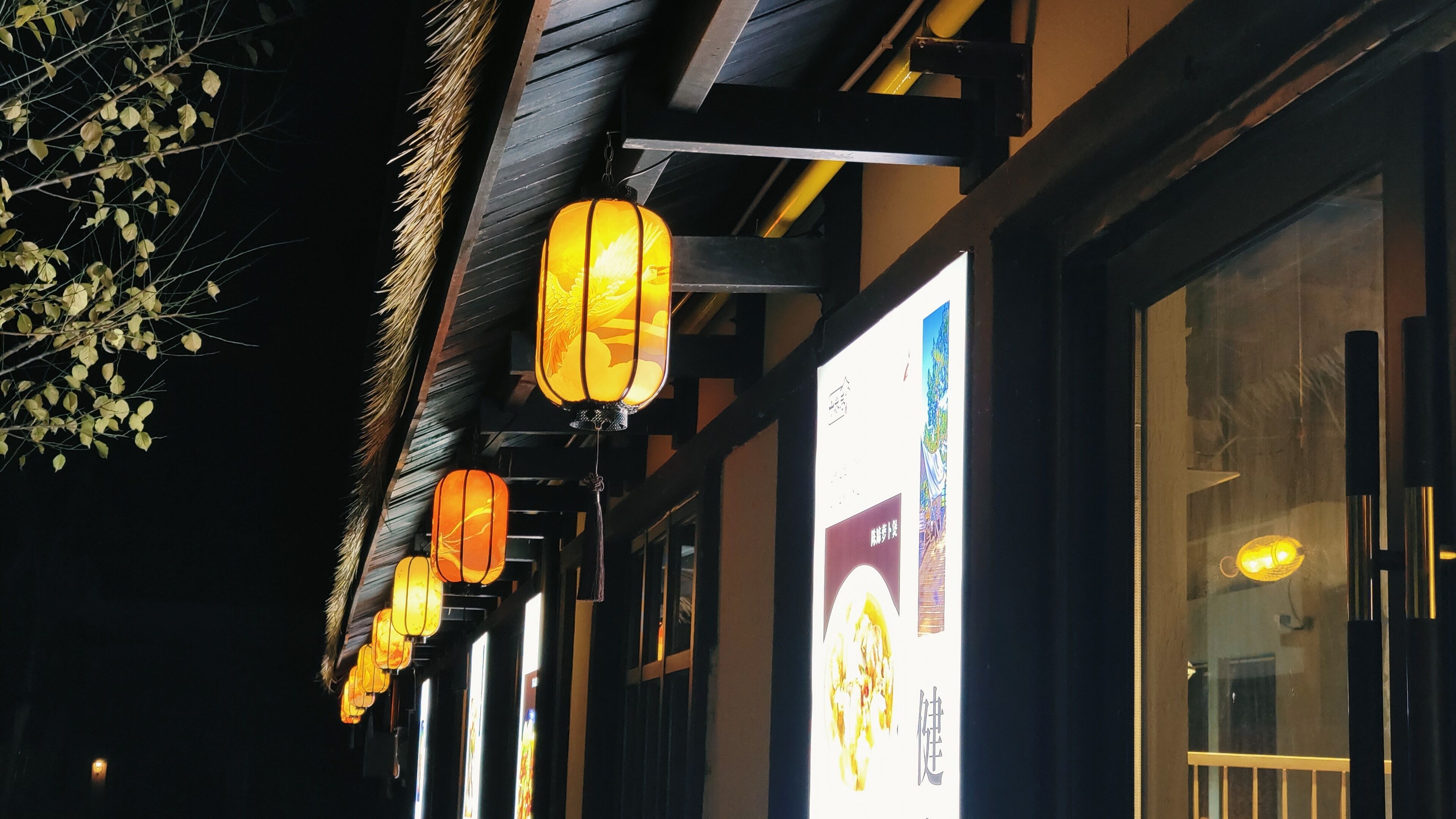 A building facade with traditional lanterns hanging from the roof, a large illuminated sign, and a window with a view of the interior.