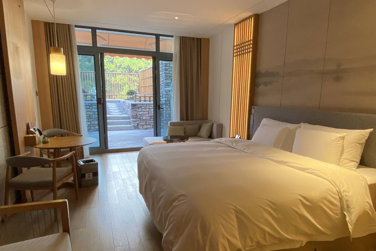 A hotel room with a large bed, a small table with chairs, a sofa, and a view of the outside through a glass door.