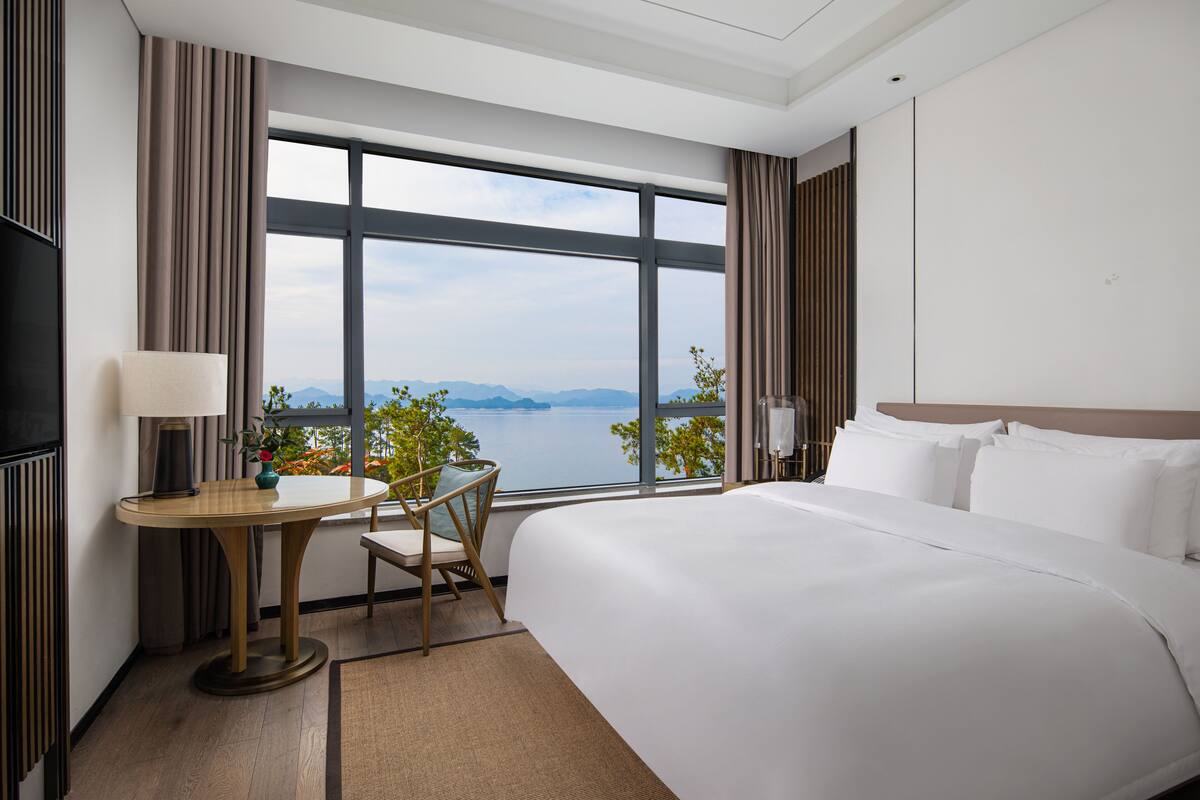 A hotel room with a large bed, a desk with a lamp, a chair, and a view of the water and mountains through large windows.