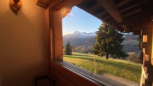 Ein Zimmer mit großem Fenster und Blick auf eine Berglandschaft, Holzbalken und einer an der Wand montierten Leuchte.