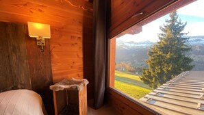 Ein Zimmer in einer Holzhütte mit einem Bett, einem kleinen Katzenhäuschen aus Holz, einer Wandlampe und einem großen Fenster mit Blick auf eine verschneite Berglandschaft.