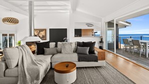Un salon moderne doté d’un grand canapé d’angle, d’une table basse ronde et offrant une vue sur l’océan grâce à de grandes portes-fenêtres coulissantes.