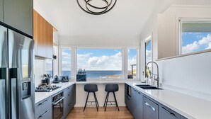 Une cuisine moderne dotée d’une grande fenêtre donnant sur la mer, d’appareils électroménagers en acier inoxydable et de deux tabourets de bar.
