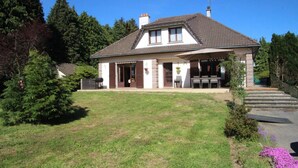 A two-story house with a covered patio, a lawn, and a small garden with purple flowers.