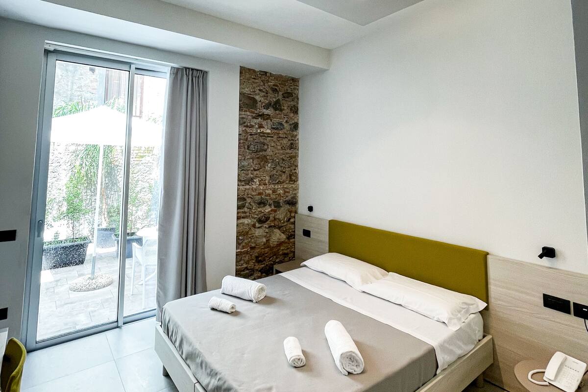 A modern hotel room with a bed, a small table with a phone, and a view of the outdoors through a sliding door.