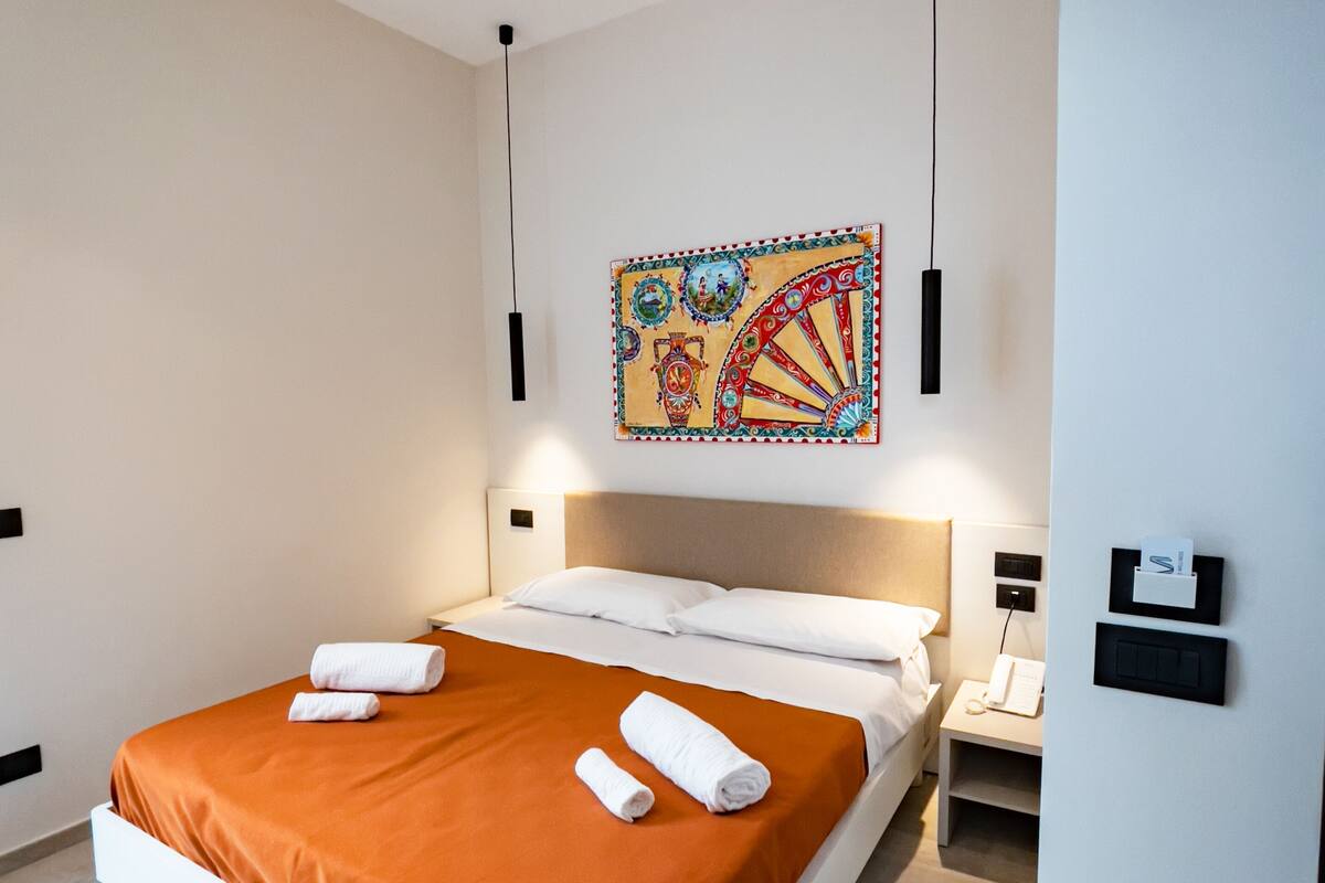 A modern hotel room with a large bed, white headboard, and a colorful artwork above the bed.