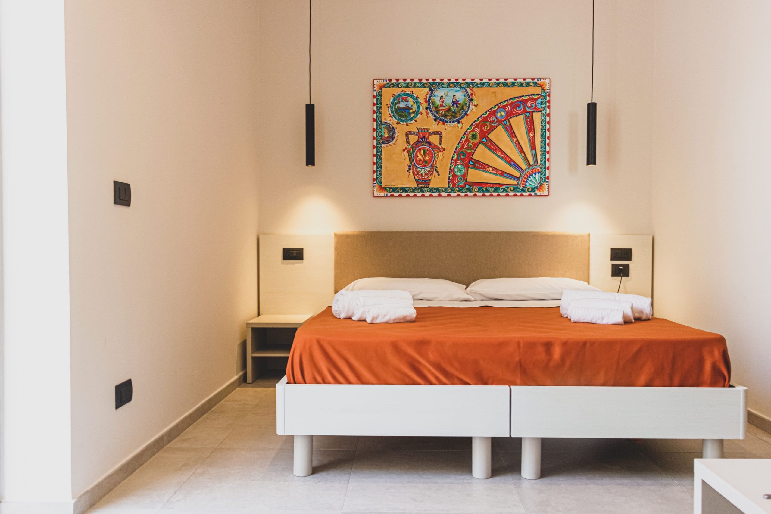 Een moderne slaapkamer met een groot bed, witte nachtkastjes en een kleurrijk wandtapijt.