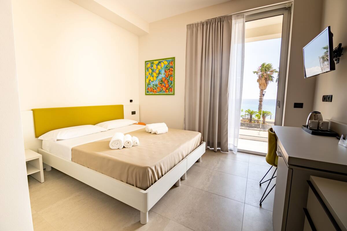 A hotel room with a bed, a desk, a TV, and a balcony with a view of the sea.
