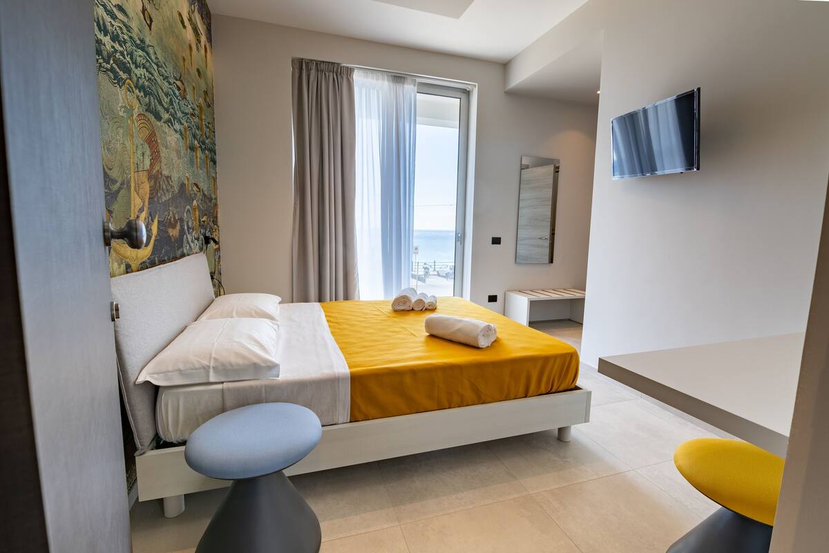 A modern hotel room with a bed, a small desk, a chair, a TV, and a view of the sea.