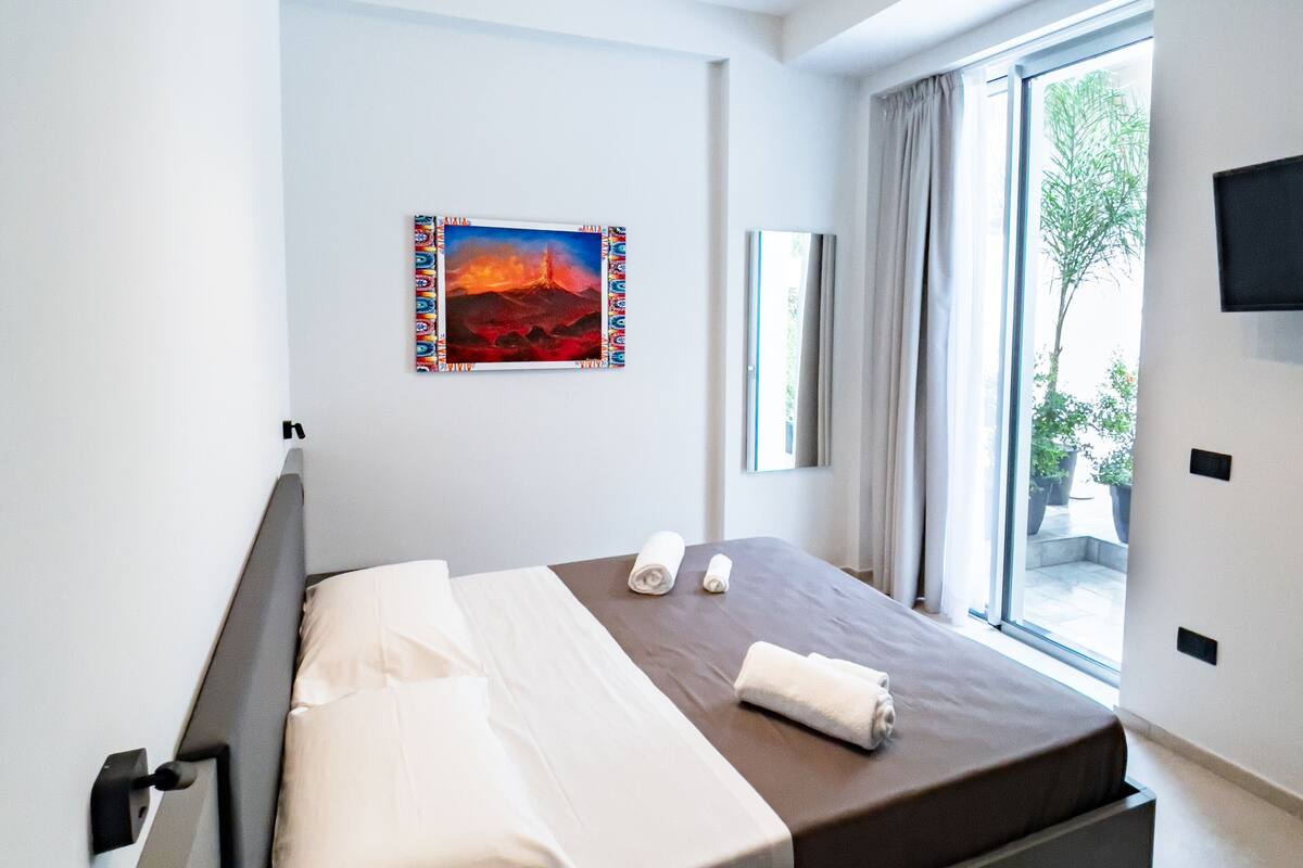 A modern hotel room with a bed, a painting, a TV, and a view of the outdoors.