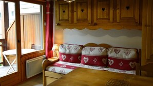 A cozy cabin interior with wooden walls, a wooden bench with heart-patterned cushions, a table with a wire rack, and a window with a view of the outside. - Cozy 1-Bedroom Apartment, Well-Equipped, Near Ski Slopes and Village Center (Crest-Voland)