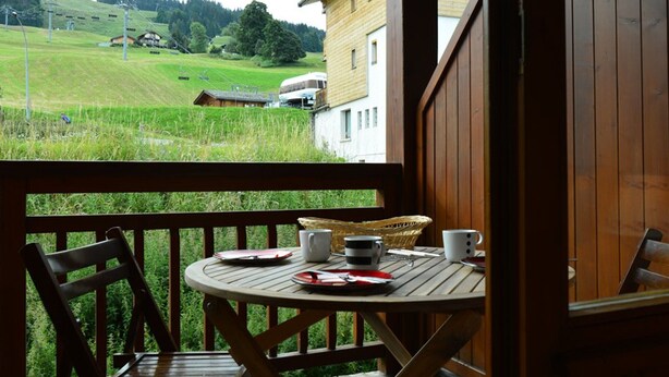 A wooden deck with a table set for two, overlooking a green landscape with buildings in the distance. - Cozy 1-Bedroom Apartment, Well-Equipped, Near Ski Slopes and Village Center (Crest-Voland)