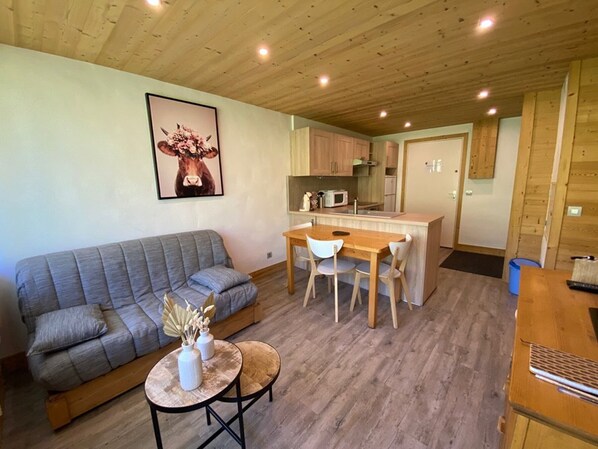 Un espacio de sala acogedor con un sofá gris, una pequeña mesa redonda con adornos, y una mesa de comedor de madera con sillas. Se ve una cocina con armarios de madera y un microondas al fondo.