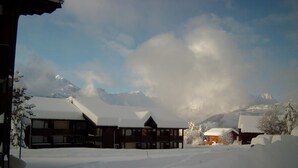 Un edificio cubierto de nieve con fachada de madera y múltiples ventanas, rodeado de árboles nevados y montañas al fondo.